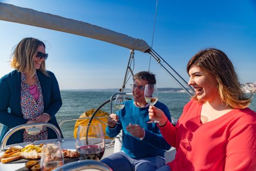 Três passageiros sorridentes a bordo de um veleiro, brindando com copos de vinho branco junto a uma tábua de aperitivos, com o Rio Tejo e a costa de Belém ao fundo.