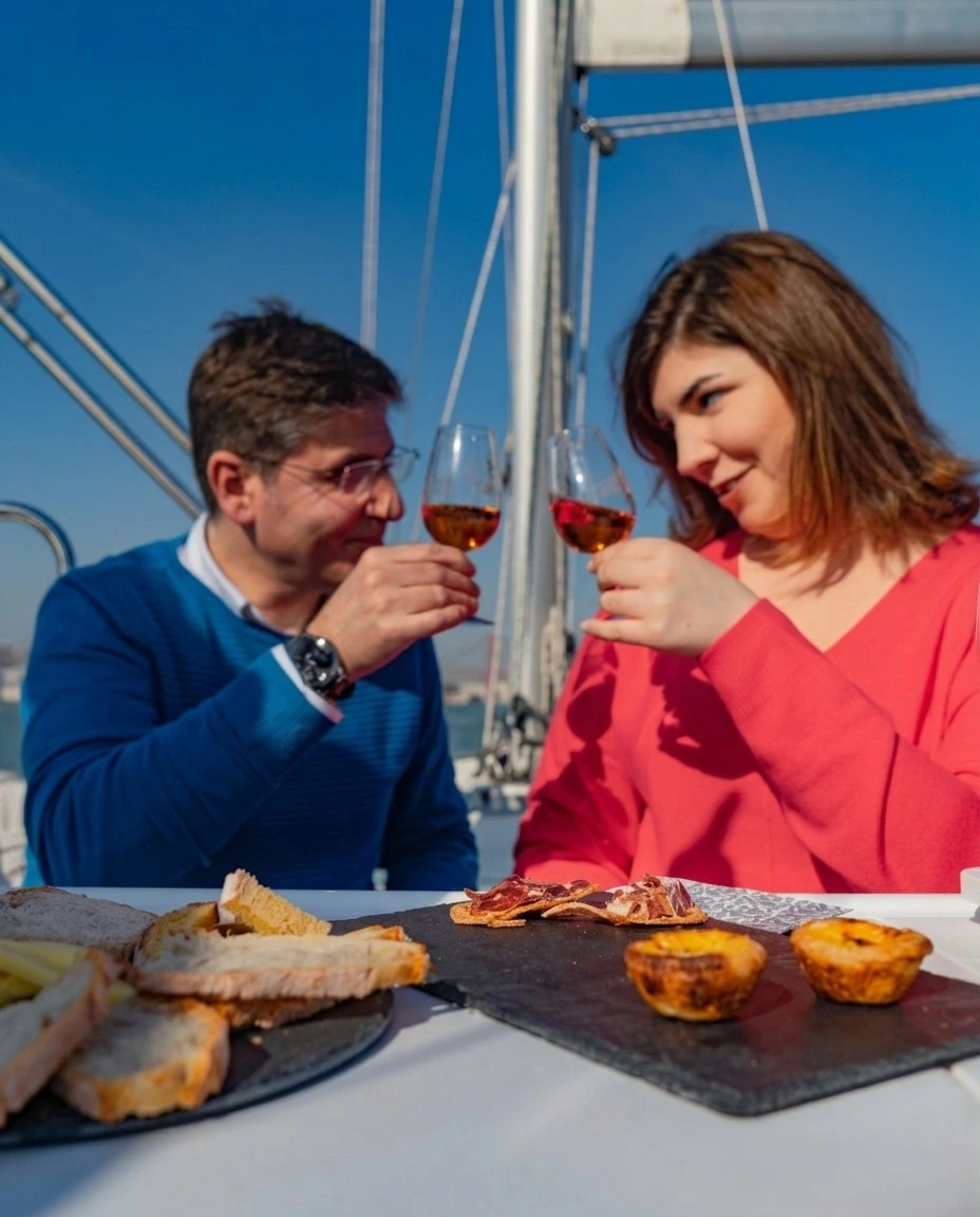 Um casal brinda durante uma prova de vinhos gourmet a bordo de um veleiro em Lisboa, com petiscos tradicionais na mesa e céu azul.