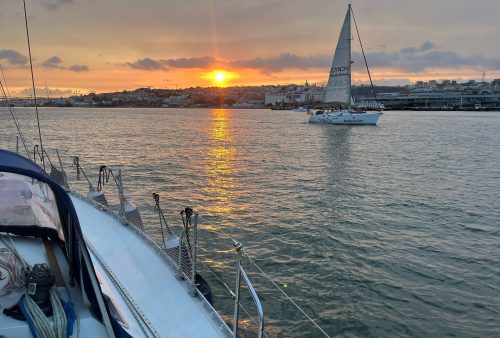 Vista a partir do convés de um veleiro no Rio Tejo durante o pôr do sol, com outro veleiro navegando e a silhueta da cidade de Lisboa ao fundo.