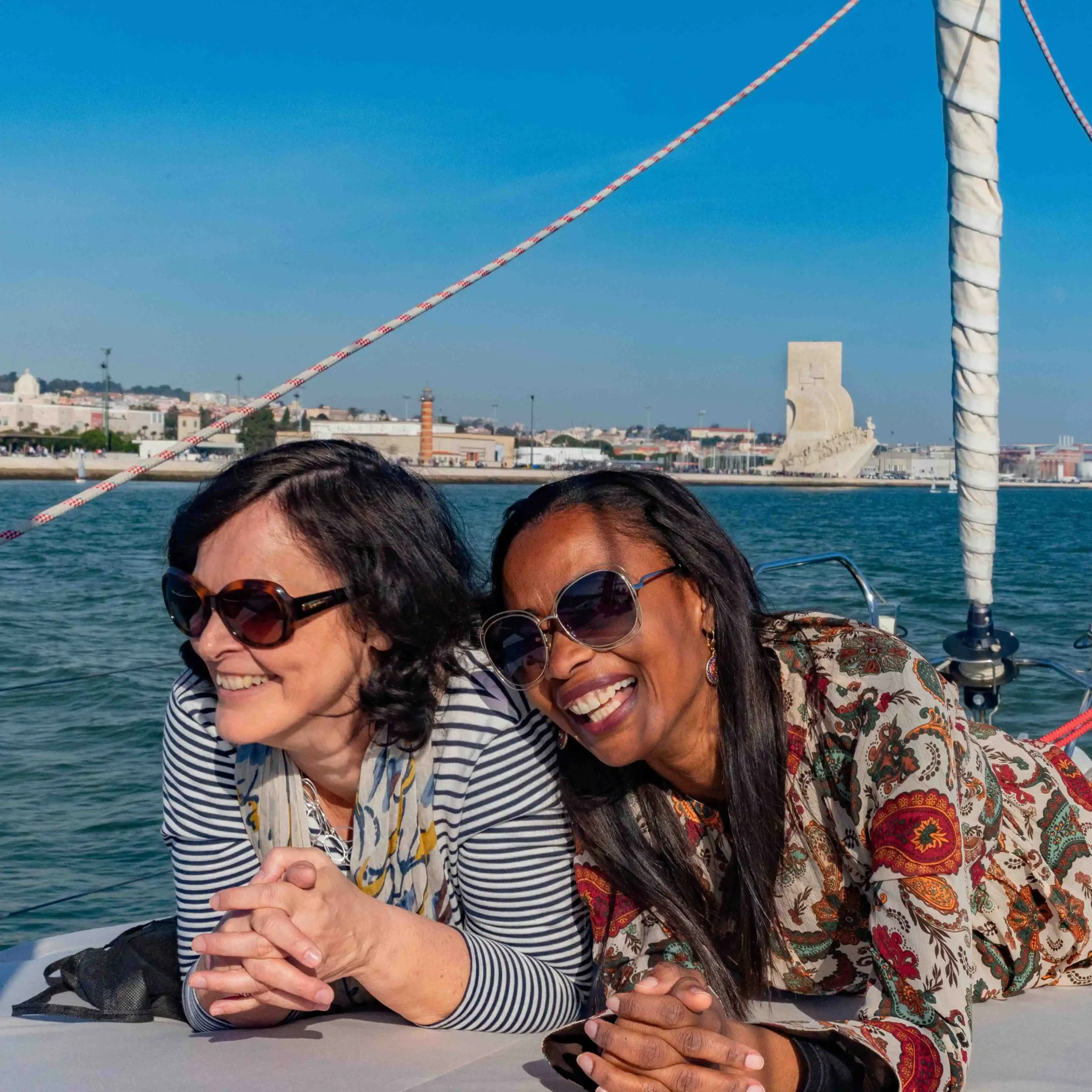 Duas mulheres sorridentes na proa de um veleiro no Rio Tejo com o Padrão dos Descobrimentos e a Ponte 25 de Abril ao fundo.