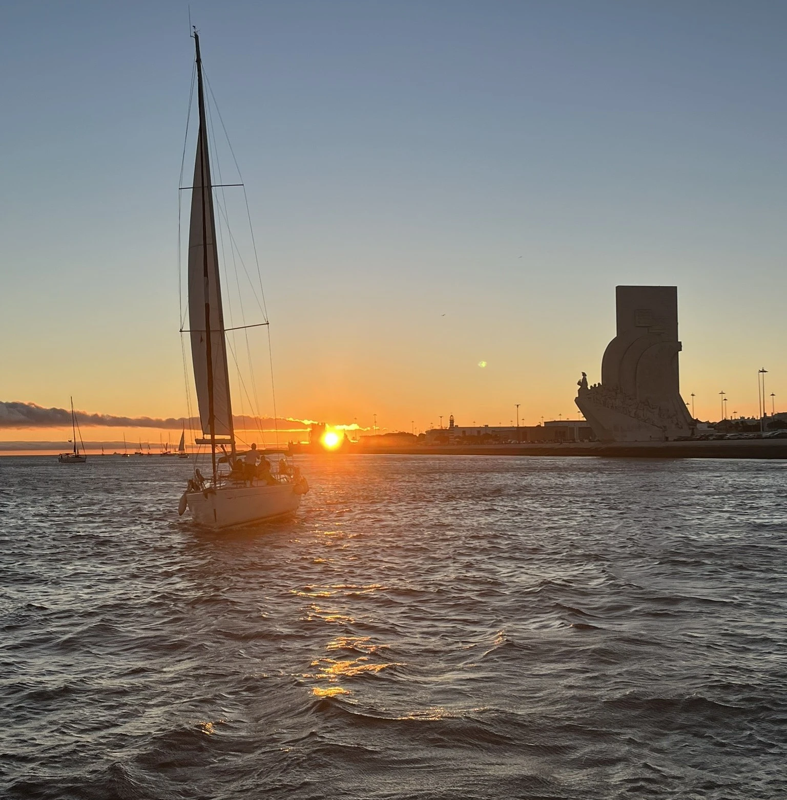 Sailboat navigating the Tagus River during sunset with the silhouette of the Padrão dos Descobrimentos in the background.