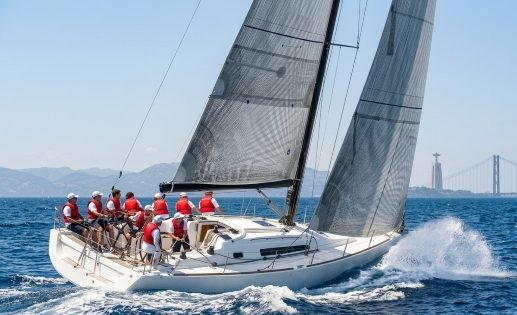 Um veleiro a navegar nas águas do Rio Tejo em Lisboa, com uma tripulação ativa de pessoas a trabalhar em conjunto nas manobras. No fundo, a Ponte 25 de Abril e o monumento ao Cristo Rei sobre um cenário de colinas distantes sob um céu azul claro.